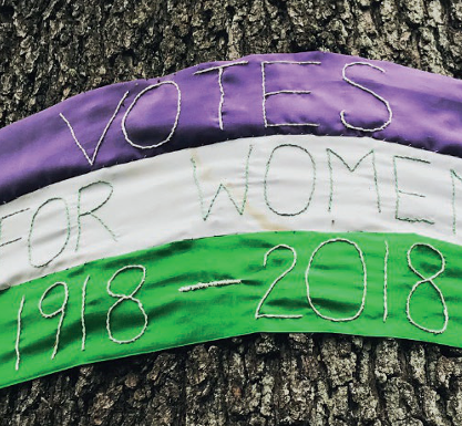 Votes for Women 1918-2018 stitched onto a banner in the suffragette colours, purble, white, green. The banner is wrapped around a tree
