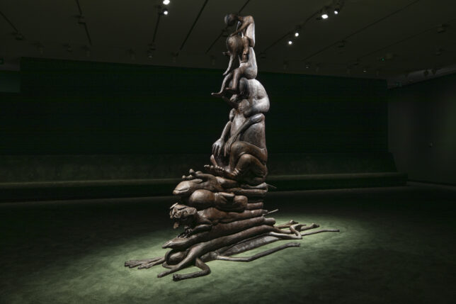 Bronze sculpture on a lush green carpet depicting several humanoid figures balanced on each other. The figures at the bottom of the pile are completely flattened whilst the top ones all have their heads hanging down creating a despondent mood.