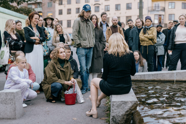 a woman with her back to the camera sits on the edge of a fountain a crown of people a standing close to her at different levels, watching.