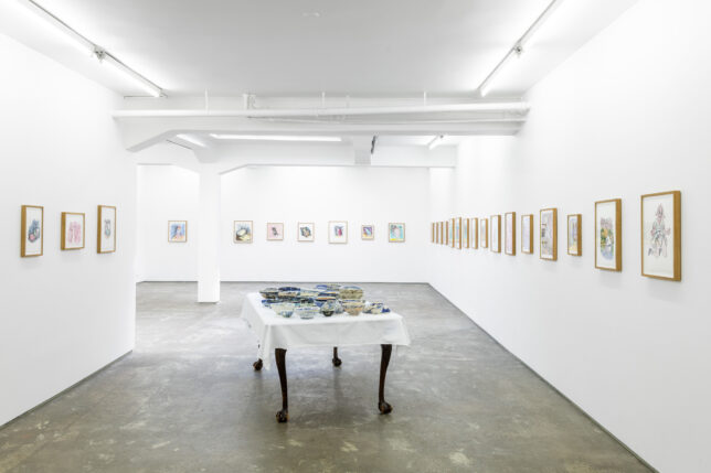 Exhibition view of a room with lots of small pictures lining the walls and a table in the centre with ceramic bowls