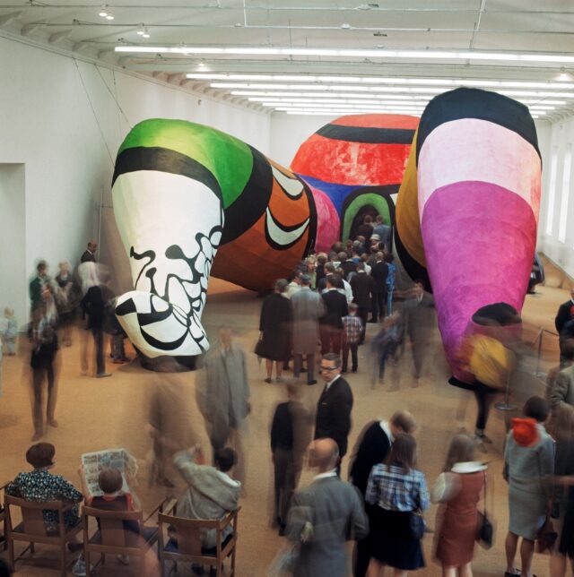 Installation shot from an exhibition in Stockholm in 1966 showing lots of participants queuing up to enter between the legs of a giant multi-coloured plaster-cast sculpture. figure in a birthing position. 