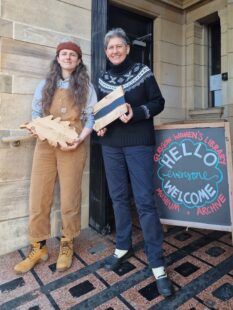 Sue and Annie stand sie by side in the entrance to GWL. They are smilling and holding an oak cheese board each.