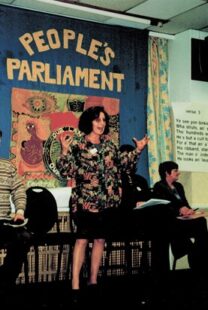 An old looking photograph of Cathy McCormack. She stands in front of a banner, her hands held out as if emphasising the words she is saying. She stands in front of a banner that says People's Parliament