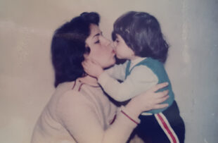 An old looking photograph shows a woman with black hair holding a child and kissing them on the lips. The child who is probably about 2 holds their hands up to the womans face