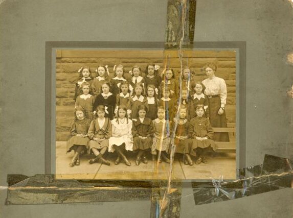 An old photograph taped into an album page with yellowing tape, of a class of girls in rows with their teacher