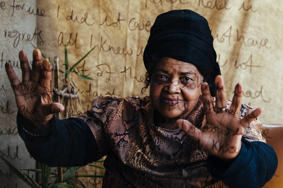 A photo of Miriam Tlali, a black woman with a black wrap on her head. Her hands are outstretched, palms facing the viewer. Overimposed on top of the image is handwriting of a famour quote from her work