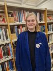 A photo of Annika, a white woman with shoulder length blonde hair. She is wearing a dark blue shirt over a blacl top and wears a badge with her name on it and another badge with her pronouns displayed 'She/her'. Annika is standing in the library in front of shelves of books