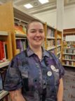 A photo of Ren standing in the library in front of shelves of books. Ren has a cropped hairstyle and a patterned shirt. They are smiling and looking at the camera