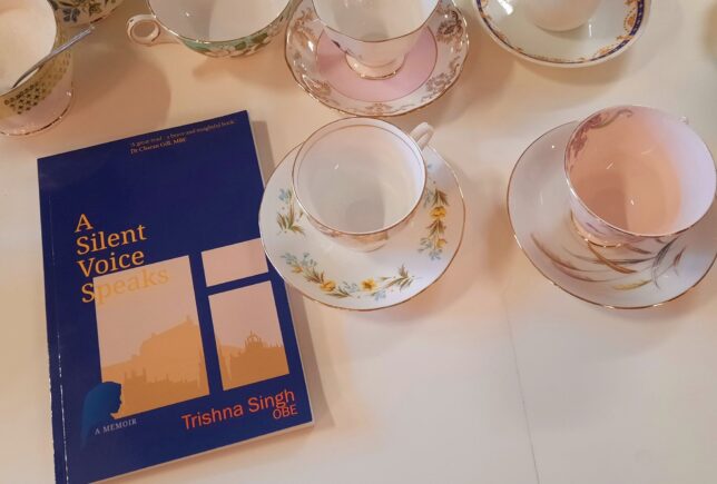 Image shows some china tea cups and saucers, with Trishna Singh's memoir, A silent voices speaks, beside them. The book has a blue cover and shows a yellow window, with a woman looking out of it.