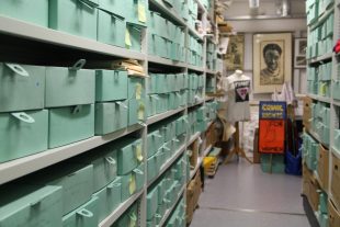 A photograph of metal shelves with green paper boxes on them. at the far end of the shelves is a mannequin with a t shirt on it and a portrait of Jackie Kay