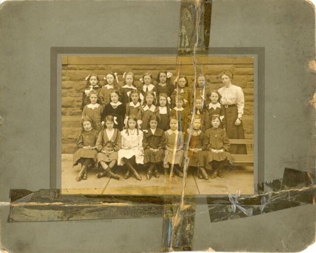 An old sepia photograph of students at Golfhill Public School, taped into an album page with yellowing tape. It is a formal school photograph with girls sitting in rows with their school mistress standing to the right.