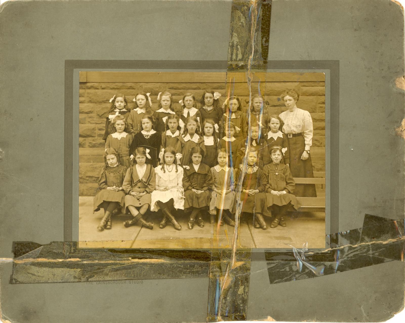 An old sepia photograph taped into an album page with yellowing tape, of a class of girls in rows with their teacher
