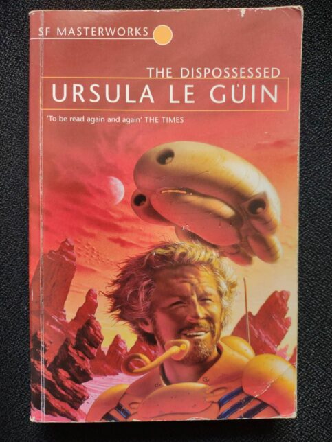 The front cover of The Dispossessed by Ursula Le Guin. It shows a smiling man under a red sky, with a spaceship behind him.