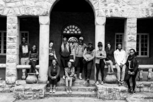 black and white image of Book Bunk team members, 12 individuals, casually dressed, smiling and informally positioned in front of a stone building with steps leading up to a porch and two central stone pillars. 
