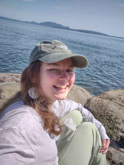 A photo of our volunteer Ashley - a white woman with long light brown hair and a grey cap on, smiling in front of the sea.