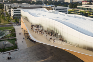 coloured image of the Oodi library building seen side on from an elevated position. The building is a rectangular shape with curved roof and large curved balcony populated with lots of visitors. 