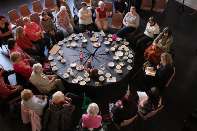 Photo taken from above. People sit round a large round table with lots of cups of tea and brightly coloured plates.