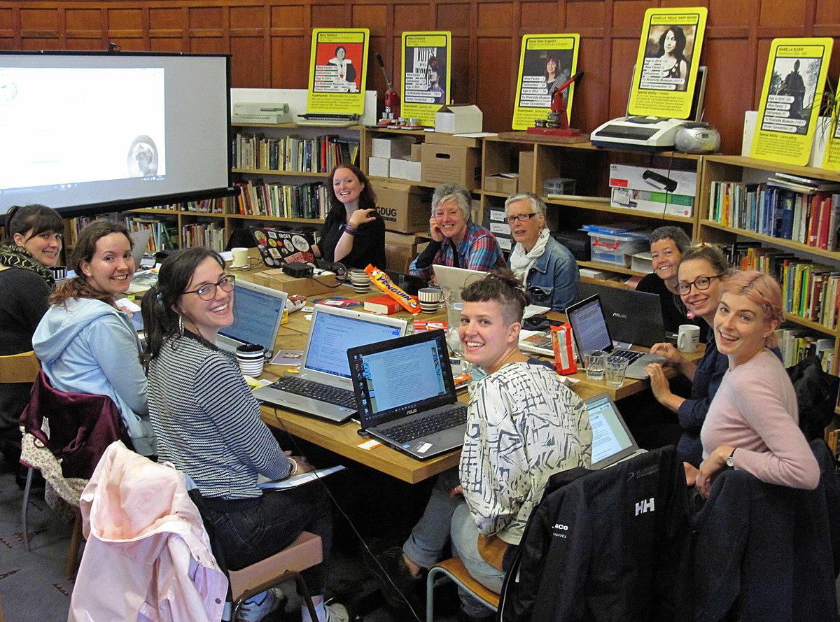 A group of people sit round a table at GWL, they are turned to face the camera, smiling and they have laptops in front of them. Behind them is a screen as well as shelves of books.