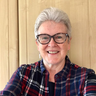 Portrait photo of a white older woman with short white hair and glasses, wearing a tartan short. She is smiling and looking into the camera.