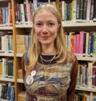 Annie Goddard, a white woman with long blonde hair stands in front of the bookshelves in the library