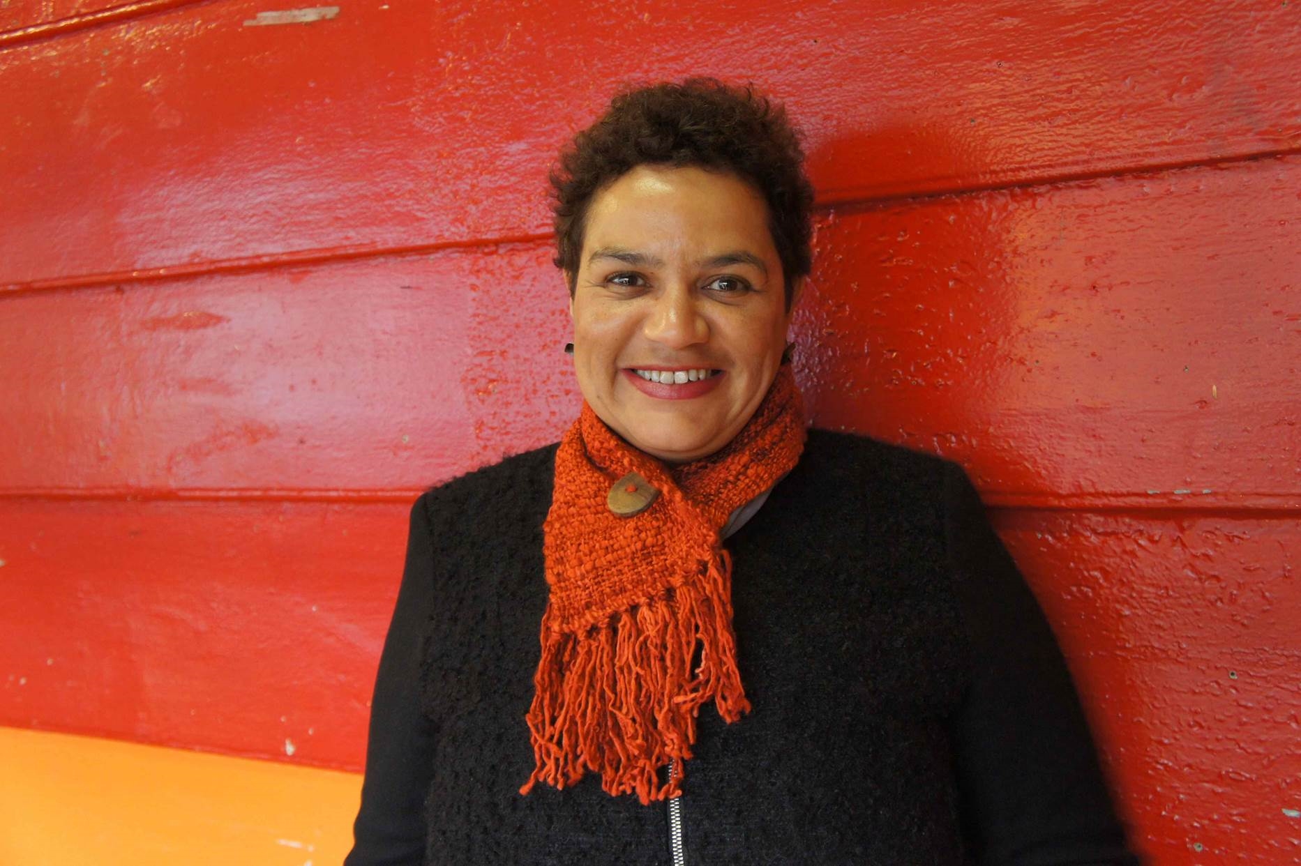 A photograph of Jackie Kay, a woman of colour with short dark hair, standing against a red wall.