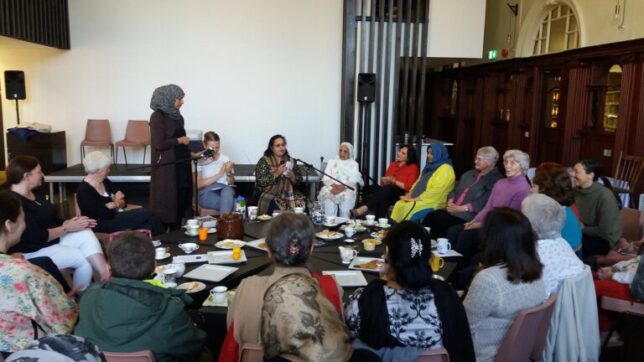 A group of women sitting around a table with cups of tea, listening to Urdu poetry read by Shamshad Ghani.