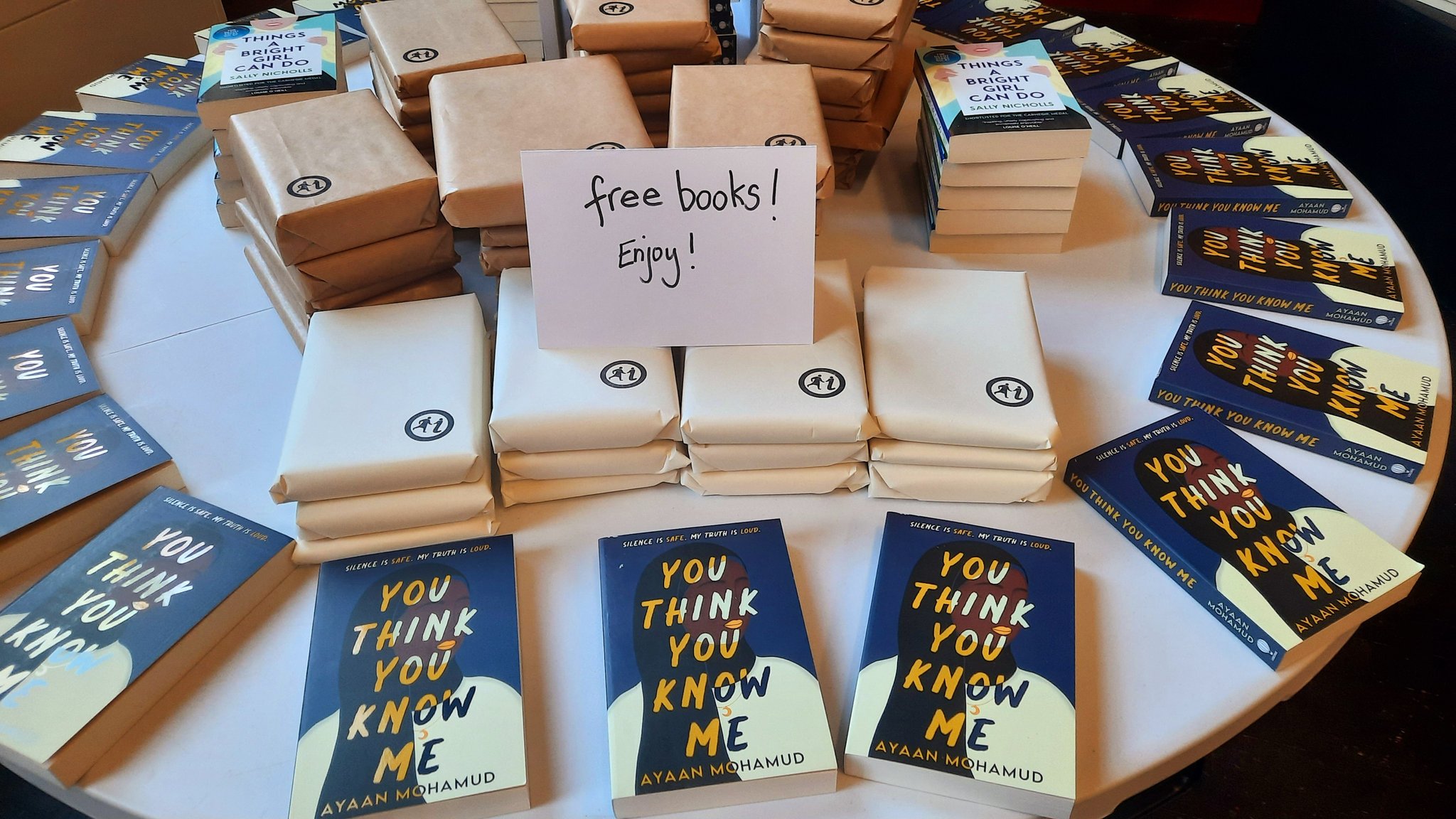 A photograph of a table with piles of books on it, some wrapped in paper, some not, and a sign saying 'Free books! Enjoy!'