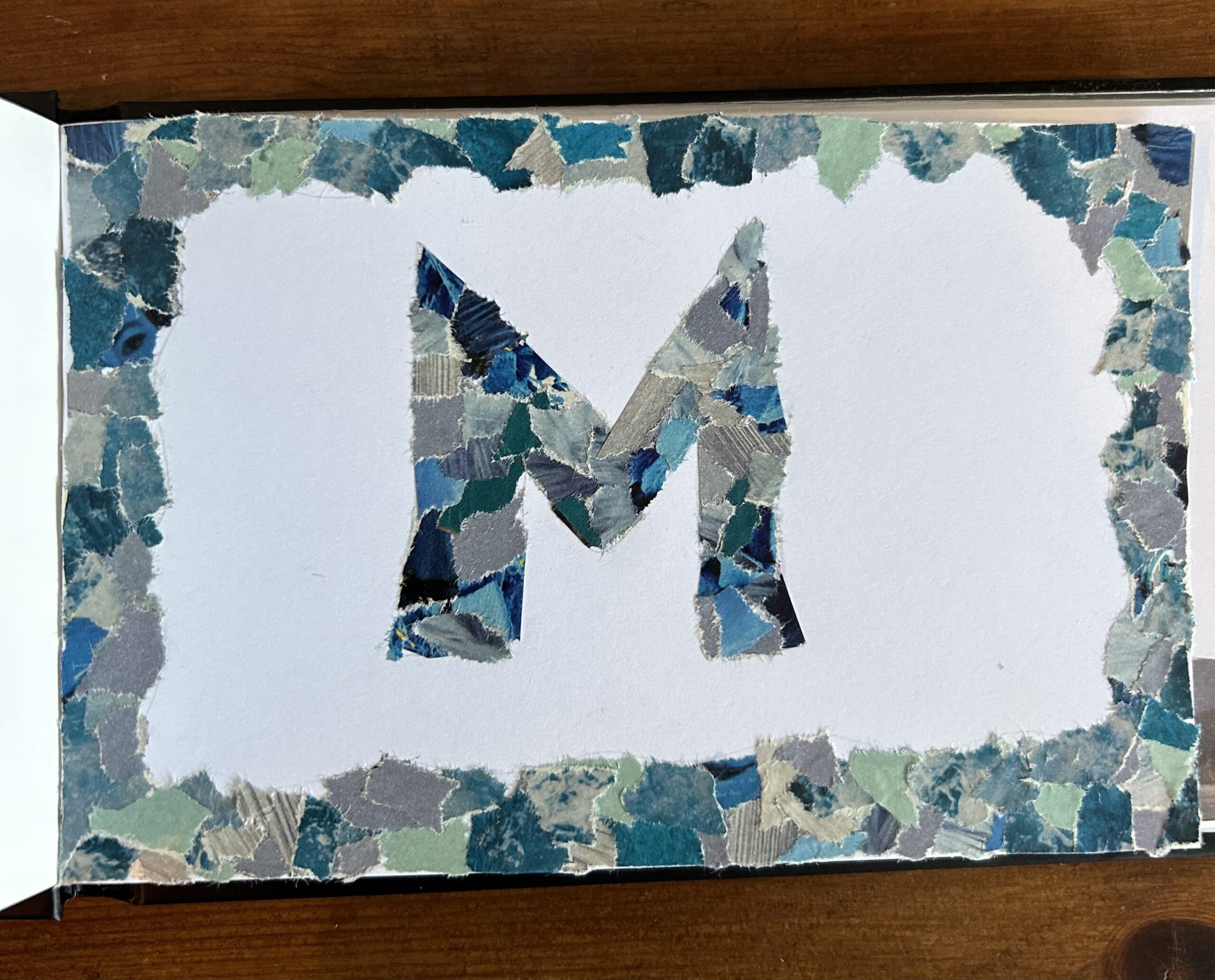 Photograph of a piece of white paper decorated with bits of blue paper collaged in the shape of the letter 'M'.