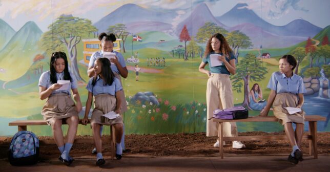 Five young women of colour stand or sit in front of a painted backdrop of a country scene, holding scripts in their hands.