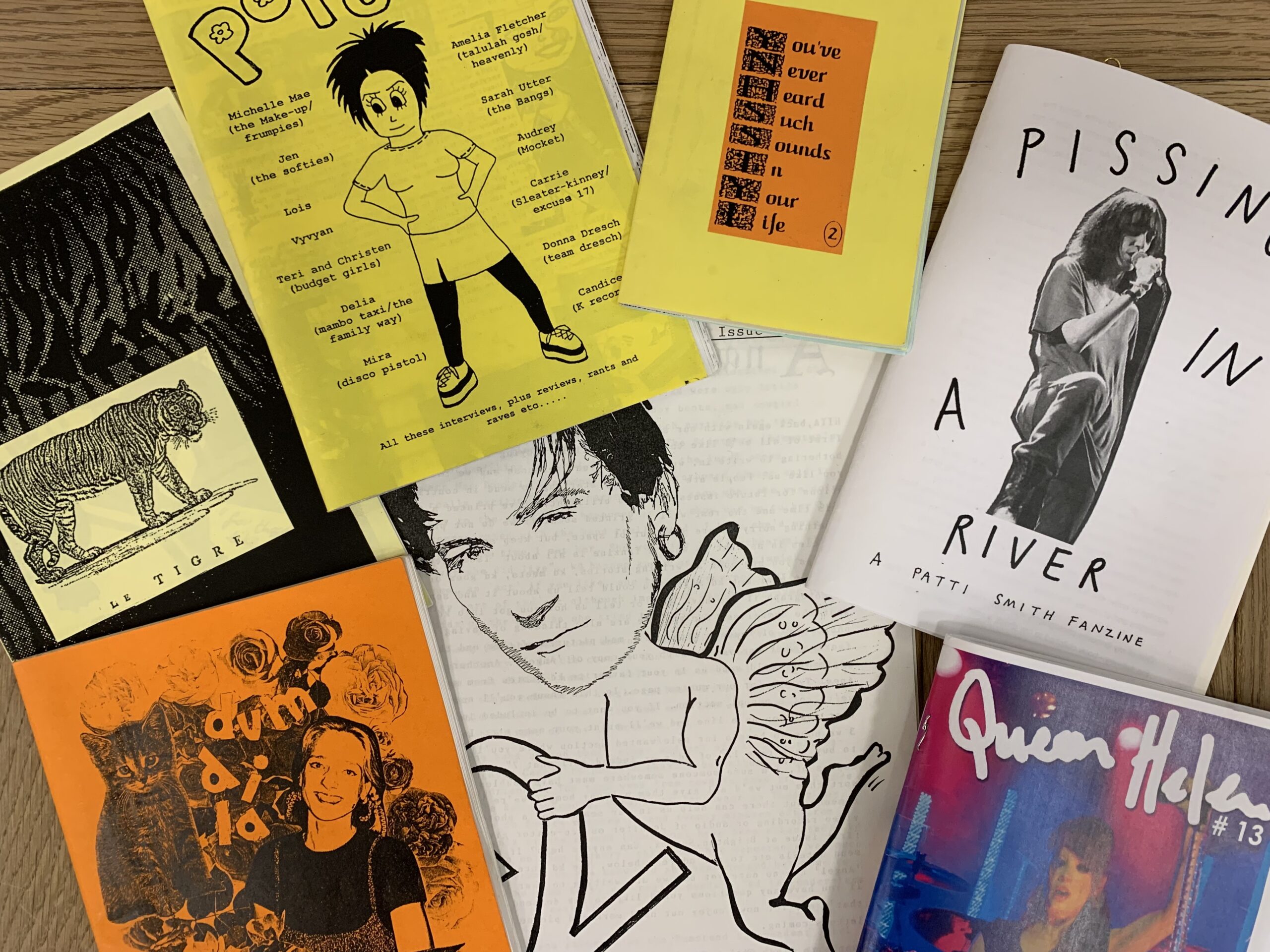A collection of music zines (handmade and self-published magazines) in colours yellow, pink, blue, orange, black and white, lying on a wooden table.