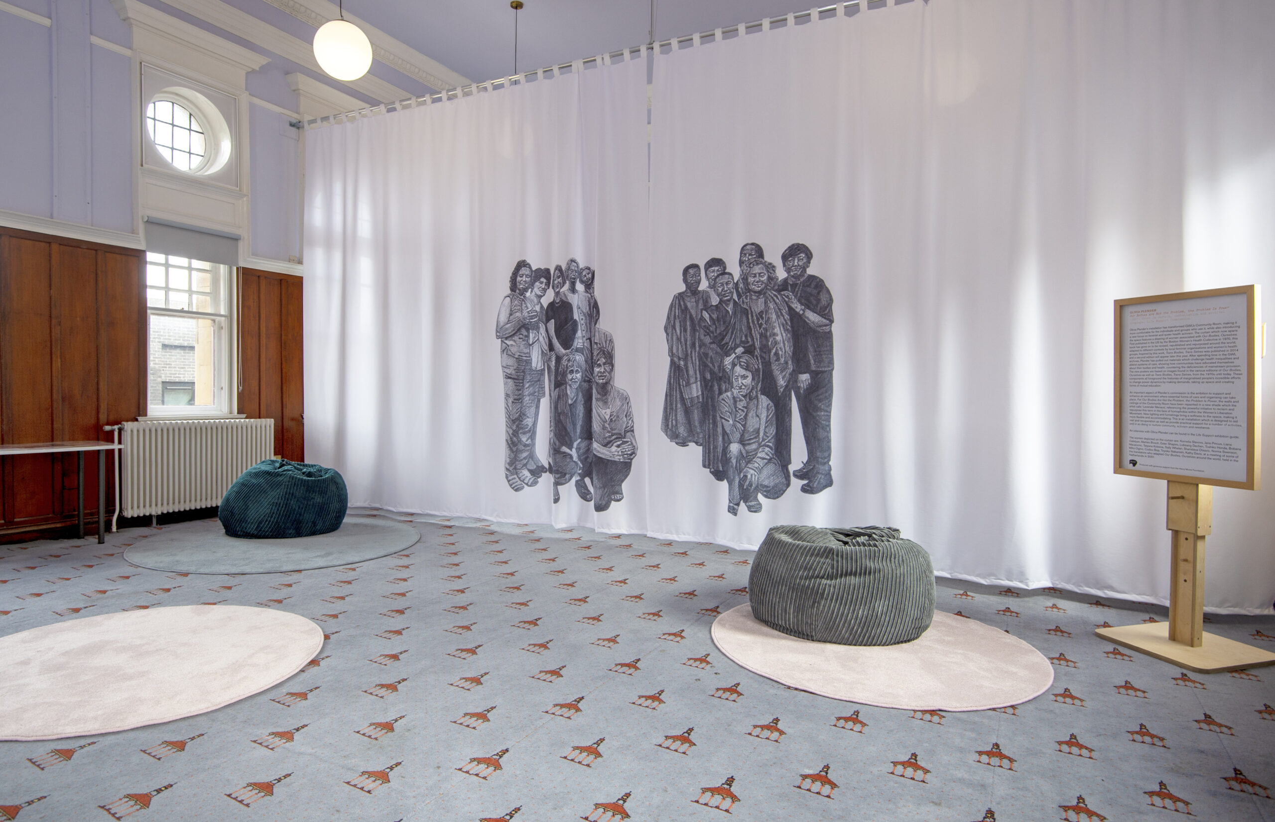 GWL's community room with a large curtain with illustrations of two groups of women printed on it. The community room is filled with soft furnishings and lavender colours.