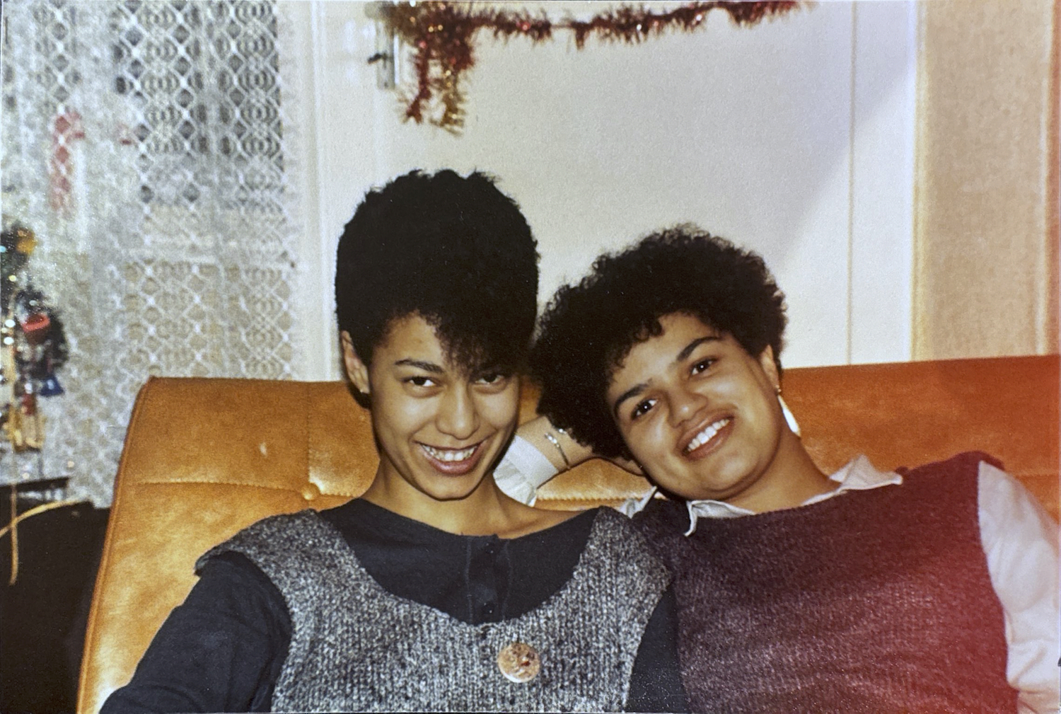 Maud Sulter and Jackie Kay sit next to each other on a sofa, probably in their early 20s, smiling. Jackie's arm is resting behind her and Maud and there is tinsel hanging behind them.