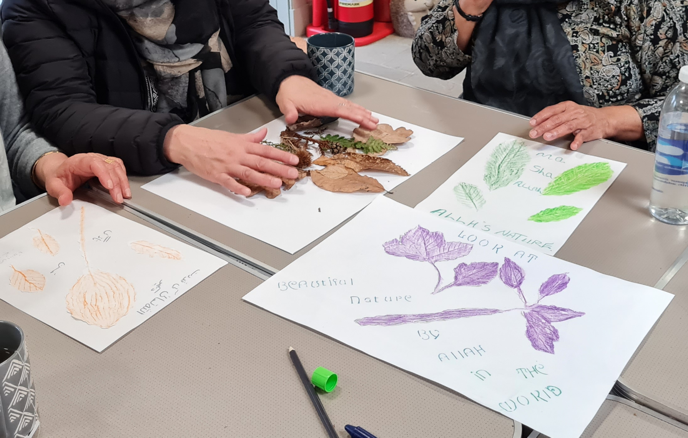 Pieces of paper are laid on a table, each with drawings of leaves surrounded by words. A collection of real leaves and twigs are laid out next to these and three people sit around the table: their hands can be seen working over the paper.