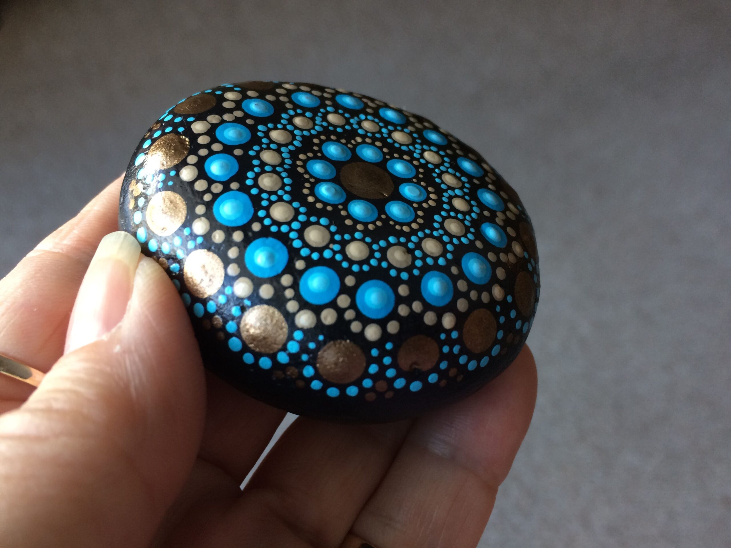 A hand holds a stone painted with a mandala pattern in blue and gold paint.