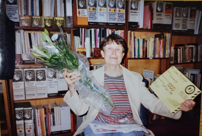 Photograph of a woman smiling and sitting in front of library shelves holding a bouquet of flowers and a certificate.