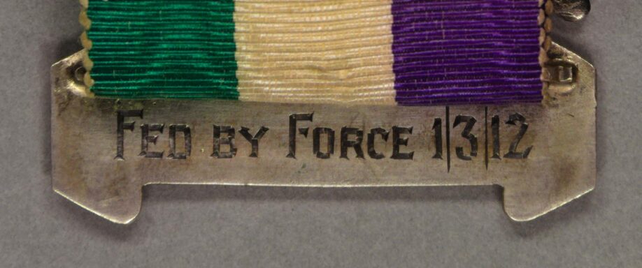 close up image of the reverse of Maud Joachim's Hunger Strike medal with the words "Fed by Force 1/3/12" inscribed on the bottom bar of the medal