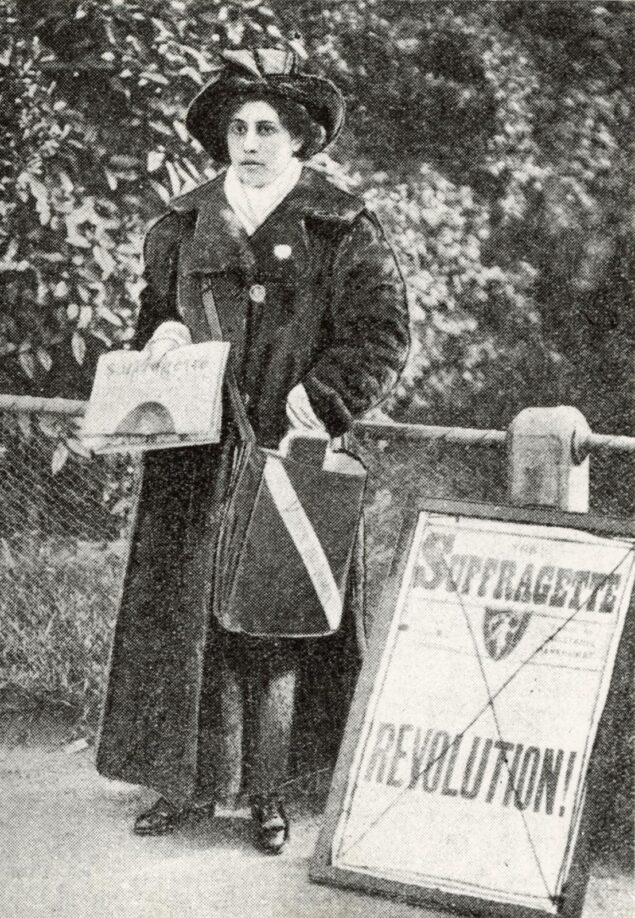 Black and white postcard of Princess Sophia Duleep Singh dressed in an overcoat and holding a copy of The Suffragette newspaper. Beside her is a newsstand advertising The Suffragette, with the word 'Revolution!' in large bold letters.