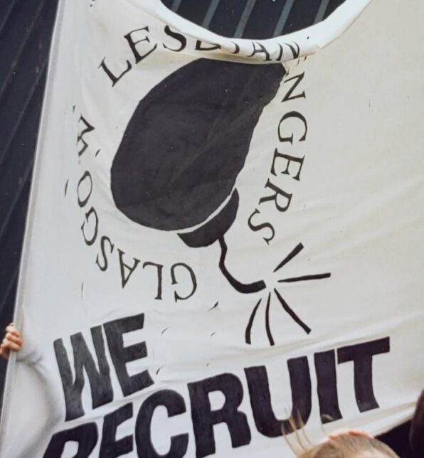 Photograph shows white banner being held up outdoors, a bomb drawing in black with the accompanying text 'Glasgow Lesbian Avengers. We Recruit'