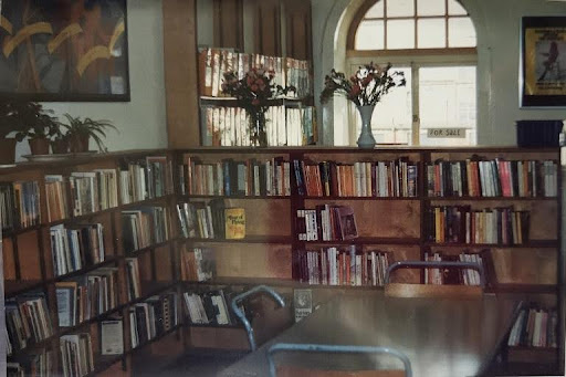 Photograph shows the inside of the library space with a table in the forefront and bookshelves surrounding table. There is a large print on the wall and the windows are large and have a distinct circular top to them.