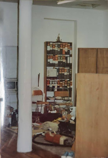 photograph showing a room used by the women's library in the 1990s. An amstrad computer on a desk, bookshelves with files on them infront of white walls. There are bags and a hoover on the floor.