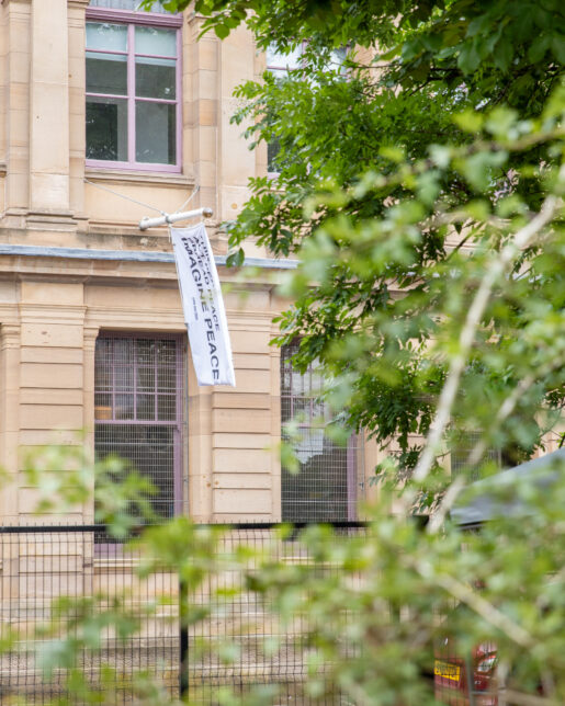 Photo shows the sandstone front of GWL with a white flag flying. the view is from the GWL garden so framed with green leaves from surrounding trees