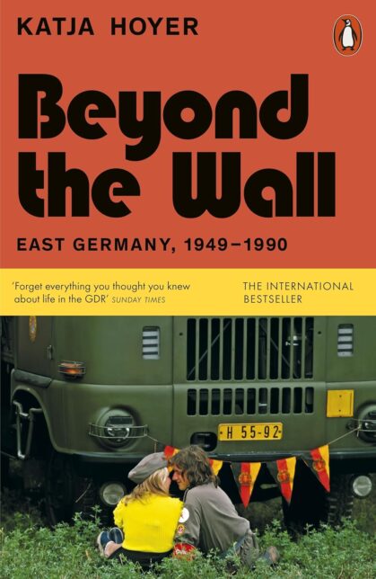 Book cover showing a young woman and a man in a uniform sitting in the grass in front of a green truck. Text reads 'Katja Hoyer - Beyond the Wall: East Germany 1949-1990 - The International Bestseller' "Forget everything you thought you knew about life in the GDR" - Sunday Times 