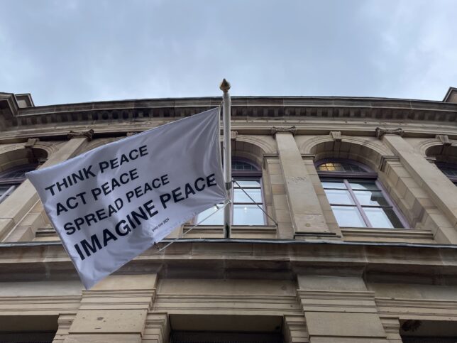 a white flag with black writing can be seen from below. It reads THINK PEACE
ACT PEACE
SPREAD PEACE
IMAGINE PEACE