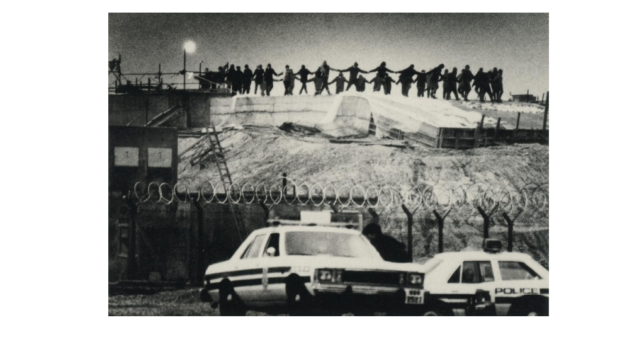 Black and white image showing silhouette's of a group raise up dancing with linked hands. In front is a barbed wire fence and police cars. 