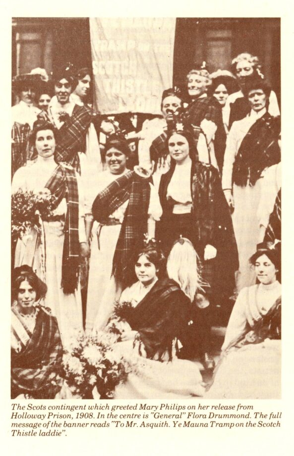 A sepia photo of Scottich suffragettes wearing white dresses and tartan sashes and shawls, holding a banner reading “To Mr Asquith. Ye Mauna Tramp on the Scotch Thistle laddie.”