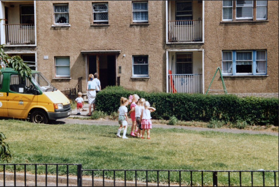 four children play on the grass in front of some flats. they are wearing summer clothes. in the background are two women with a child brushing the flat steps with a broom