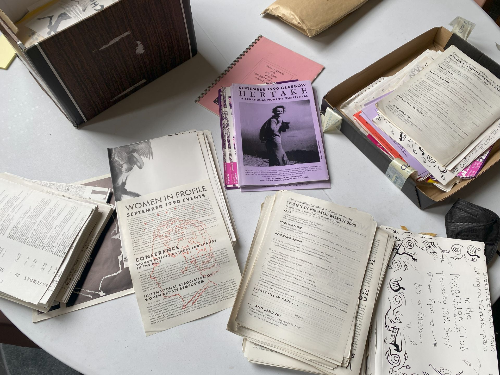 Stacks of archive material including A4 papers and large bankers boxes, folded posters, envelopes and a covid mask sit on a white circular table.