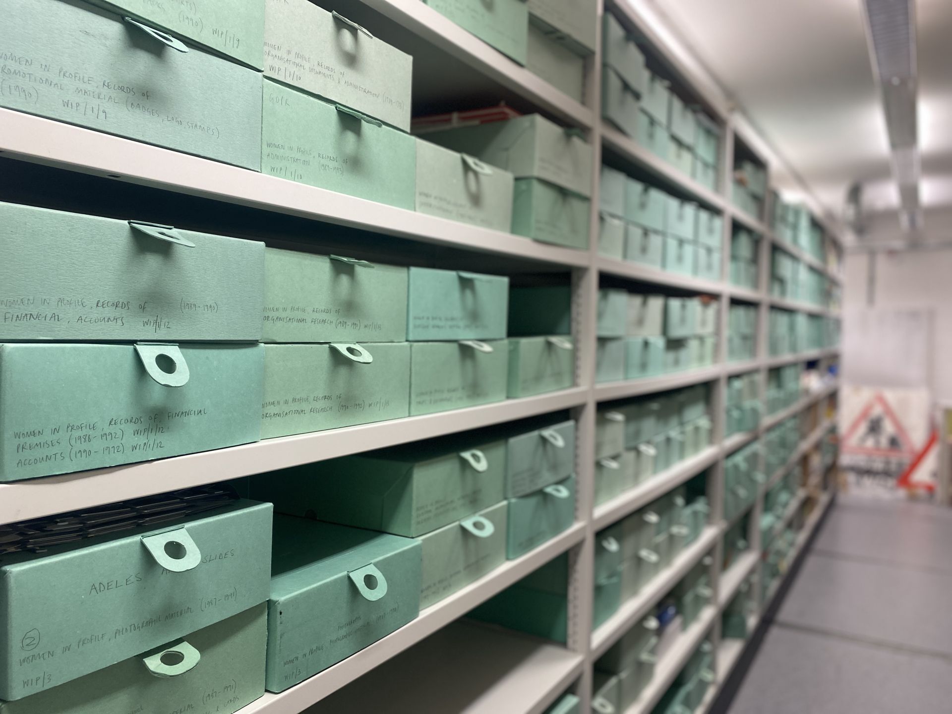 Nearly one hundred pale green boxes sit on grey metal shelves in the archive stores. In the background is a ‘women working’ sign shaped like a triangular red-rimmed road sign