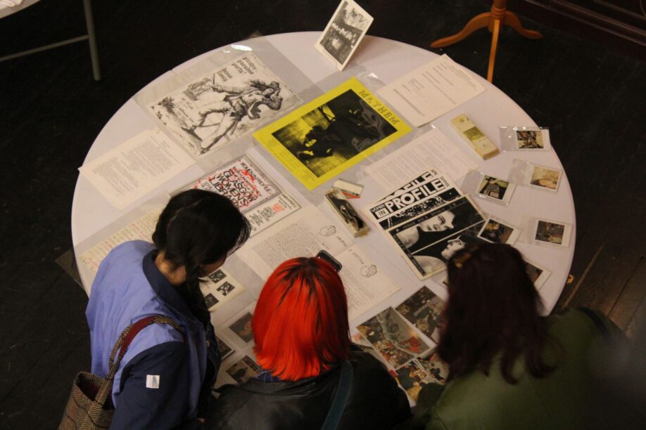 Seen from above, three people look at archive material from the GWL organisation records including posters, pamphlets, magazines and photographs, laid out on a white circular table.