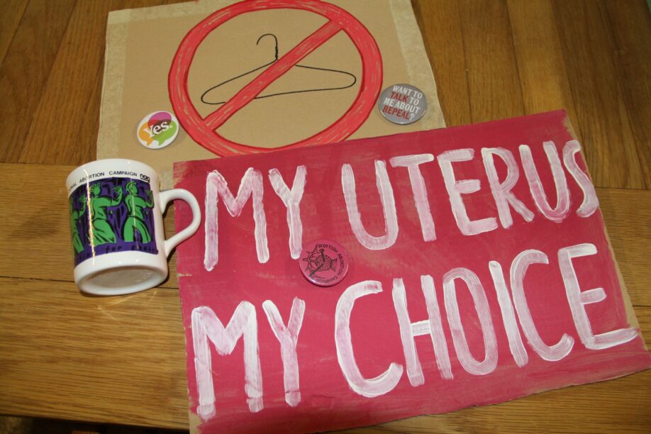 Different batches, a mug and placards for abortion rights spread on table. One placard is red and says in a white font "My uterus, my choice" and the other one displays a crossed out coat hanger.